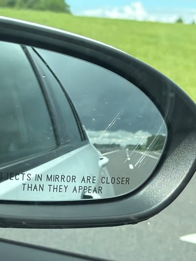 The image is a close-up, angled shot of a car's side-view mirror. The mirror is reflecting a cloudy sky, a road, and a grassy field with trees in the distance. The text "OBJECTS IN MIRROR ARE CLOSER THAN THEY APPEAR" is visible on the lower portion of the mirror. There are several water droplets on the mirror's surface and some faint scratches. The exterior of the mirror housing is black and textured. The scene appears to be outdoors, possibly on a highway, given the road reflection. The location is stated to be in the Пяршайскі сельскі Савет, Belarus, though this context is not directly discernible from the visual elements of the image itself. There are no people or explicit actions depicted in the image.