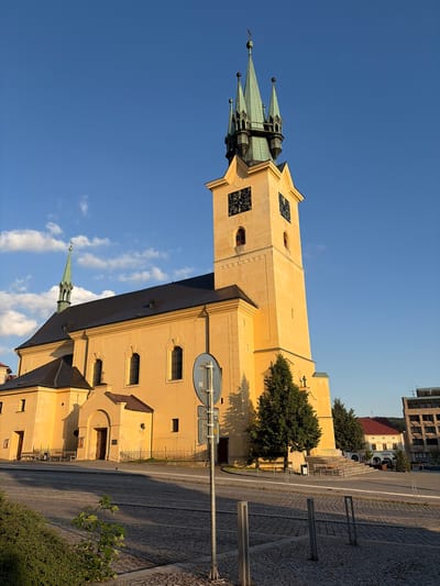 This image showcases the Saint James' Church in Příbram, Czechia, bathed in the warm light of a setting sun. The imposing yellow church features a tall, prominent bell tower with a green, multi-spired roof and clock faces. Adjacent to the tower is the main body of the church with a dark roof and arched windows. The foreground is a cobblestone plaza with benches, a fence, and a street sign. A small tree grows in the lower left corner. In the background, other buildings, including a multi-story structure and a building with a red roof, are visible. The sky is a vibrant blue with scattered white clouds. There are no people or animals visible in the frame.
