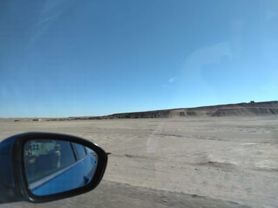 The media file shows a view from inside a car, looking out towards a vast, dry, and barren landscape under a clear blue sky. A side-view mirror in the foreground reflects the interior of the car, including what appears to be passengers and possibly a camera or phone mounted on the dashboard. The landscape itself is mostly flat, sandy, and appears arid, with a distant low ridge or cliff line forming the horizon. There are a few structures visible in the far distance, possibly buildings or industrial equipment, suggesting a remote, possibly desert or steppe environment. The time of day appears to be daytime, with bright sunlight casting minimal shadows, indicating the sun is high in the sky. The weather seems clear and dry. The scene evokes a sense of travel through a remote and undeveloped area, with a feeling of openness and isolation. No text is visible in the image.