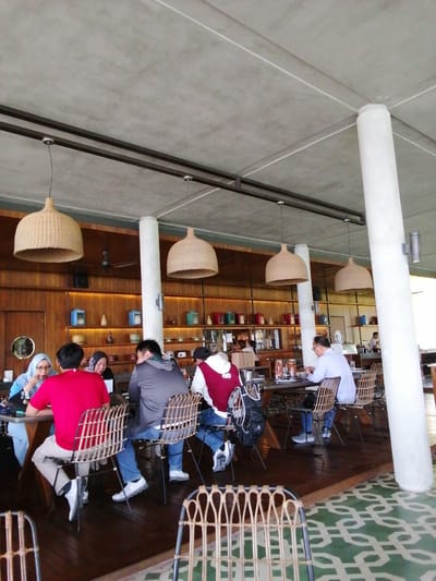 A group of people are seated at tables in a modern cafe or restaurant in Banyuwangi, Indonesia. The cafe has a high, unfinished concrete ceiling with exposed beams and large, woven pendant lights. White cylindrical pillars support the ceiling. The walls are paneled with warm-toned wood and feature shelves displaying various decorative items and containers. The flooring is dark wood with a distinctive green and white geometric pattern.
At the foreground table, a person in a bright red shirt is seated, facing away from the camera. To their left, a person wearing a light blue hijab is also seated, looking down at something on the table. Another person wearing a dark grey hoodie is seated to the right of the person in red.
In the middle ground, several other individuals are seated at tables. One person in a light blue shirt is looking at their phone, while others are engaged in conversation or looking at their meals. The tables are set with plates and drinks. In the background, more seating and tables are visible, and a server can be seen behind a counter. The overall atmosphere appears relaxed and casual.
