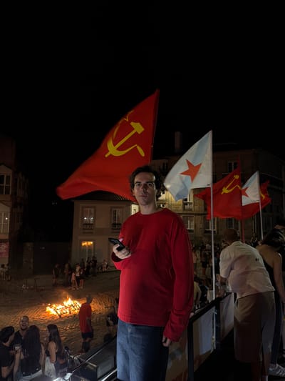 This media file depicts an outdoor gathering at night in what appears to be a city square in Santiago de Compostela, Spain. The central subject is a person with dark, curly hair wearing a red long-sleeved shirt and dark jeans, holding a smartphone in their right hand. They are standing in front of several flags, including a prominent red flag with a yellow hammer and sickle symbol, a symbol of communism. To the left of this flag is another red flag with a white star, and to the left of that, a white flag with a red star.
The background shows buildings with windows, suggesting an urban environment. A bonfire is visible in the middle ground, with several people gathered around it. Other individuals are scattered throughout the scene, some appearing to be spectators. The overall atmosphere seems festive or celebratory, given the presence of the bonfire and the gathering of people. The lighting suggests it is nighttime. There is no visible text in the image.