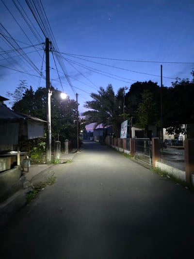 The image depicts a street scene at dusk in the city of Bireuenmeunasahblang, Indonesia. A paved road runs through the center of the frame, illuminated by streetlights. On either side of the road are houses and fences, with vegetation and trees present. Overhead, a complex network of electrical wires is visible against the twilight sky, which transitions from a deep blue to lighter hues near the horizon. A single, bright star is visible in the upper right quadrant of the sky. The scene appears quiet and deserted.