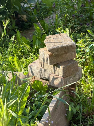 A stack of four weathered, light brown bricks is arranged in a stepped pyramid on a wooden structure in a lush, sunlit outdoor environment. The bricks are approximately rectangular with visible textured surfaces. The wooden structure appears to be a simple, rustic construction, possibly part of a garden or yard feature. The surrounding area is overgrown with various green plants and grasses, some with small purple flowers. The sunlight casts distinct shadows on the bricks and the surrounding foliage, indicating a sunny day. The background is filled with more greenery, suggesting a natural or garden setting within the city of Petukhi, Russia. There are no people or animals visible in the image.