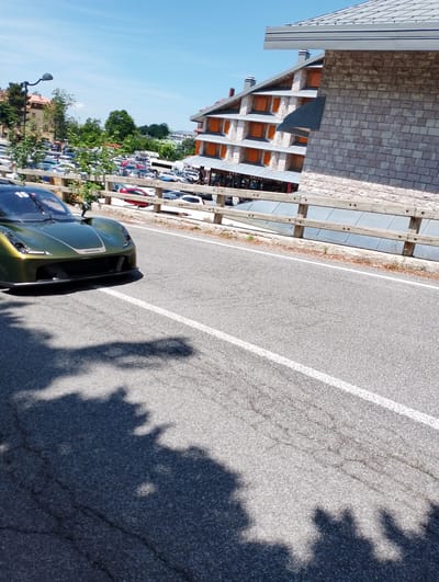 An olive-green sports car is parked on an asphalt road. The car has the number 15 on its windshield. In the background, there is a wooden fence, a parking lot with many cars, a multi-story building with orange windows, and a clear blue sky. The shadows of trees are cast on the road in front of the car.