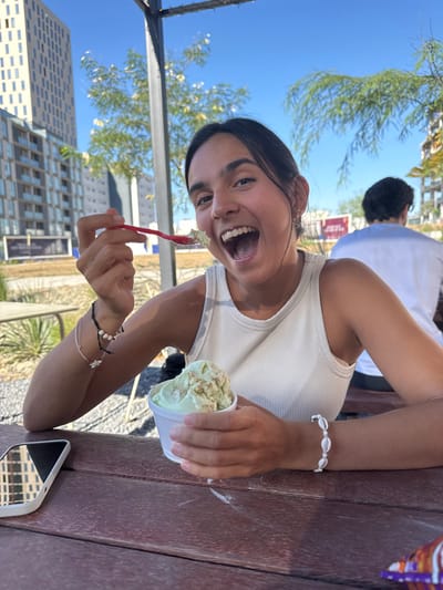 A young woman, wearing a white tank top and a white shell bracelet, sits at a wooden table in a park, laughing as she eats green ice cream with a red plastic spoon. The table is partially obscured by a metal bar, which is likely part of a shade structure. There is a cell phone on the table and a plastic bag on the right side of the frame. Another person, wearing a white t-shirt, is partially visible sitting at the table behind the woman. The woman is positioned in the center of the image, facing the camera. The scene is located in Ciudad Santa Catarina, Mexico, at the coordinates 25.658 latitude, -100.438 longitude. The location is likely a park or outdoor area. The buildings in the background suggest an urban setting. The sunny, cloudless sky suggests it is a warm day.