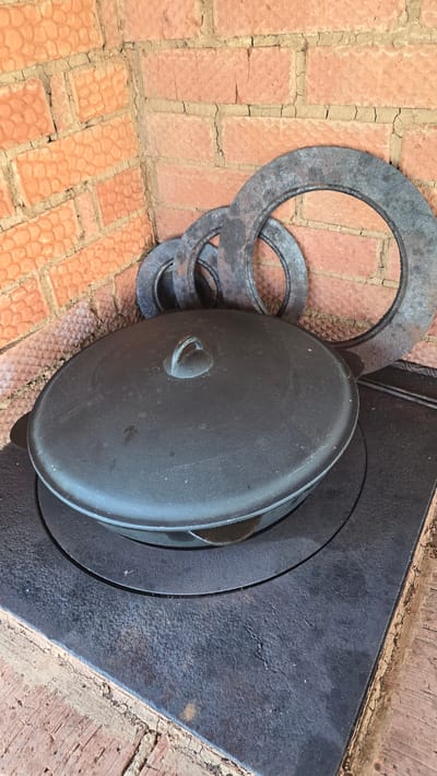 The image depicts a cast-iron cooking setup situated in Забегалово, Russia. A large, dark grey cast-iron pot with a fitted lid and a central loop handle rests on a black, rectangular cast-iron stovetop. The stovetop features a circular indentation where the pot is seated, suggesting it's part of a traditional cooker or wood-burning stove. Behind the stovetop, three concentric, dark metallic rings of varying diameters lean against a rustic brick wall. The wall consists of reddish-orange bricks with textured surfaces and irregular, cracked mortar joints, forming an interior corner. No individuals or active processes are visible in the scene.