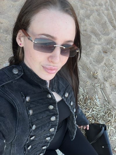 A female subject is depicted in a medium-shot, from a slightly elevated perspective, seated on a sandy surface identified as a beach in Repino, Russia. The subject has long, dark brown hair and fair skin with visible freckles on the forehead and cheeks. She is wearing dark-tinted, rimless sunglasses with reddish-brown, possibly wooden, temples that reflect a body of water and a distant horizon. On her visible left ear, she wears a small pink stud earring and a thin, golden-toned hoop earring. She is dressed in a dark grey or black denim jacket featuring a military-style design with multiple horizontal straps and numerous silver-toned, embossed buttons across the chest, shoulders, and cuffs. A black top is visible underneath the jacket, and she is wearing black leggings or tight-fitting trousers. A black, structured shoulder bag with a silver-toned stud is positioned beside her right leg. The ground is composed of light-colored sand with discernible ripples and is littered with scattered dried, light-colored reeds and plant debris, particularly on the right side of the frame. The subject is looking directly towards the camera with a subtle smile.