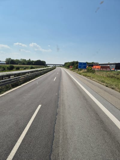 The image shows a highway scene in Germany on a clear, sunny day. The perspective is from a vehicle moving forward on a multi-lane highway. The road has asphalt surfacing with distinct white lane markings. To the left of the road is a metal guardrail separating the highway from a grassy area and a field with some trees in the distance. Further in the distance on the left, there are buildings, possibly agricultural or industrial. On the right side of the highway, there is a grassy embankment with some vegetation. A blue highway sign is visible on the right, indicating "P Bayerwaldblick -Nord 500 m". Beyond the sign, there is a building with stacked orange materials in front, suggesting a commercial or industrial area. The sky is blue with a few scattered clouds. The overall scene depicts a typical highway environment. Based on the sign, the location is near Plattling, Germany, specifically related to the "Bayerwaldblick - Nord" rest stop or viewpoint, which is 500 meters ahead. There are no people or animals visible in the image.