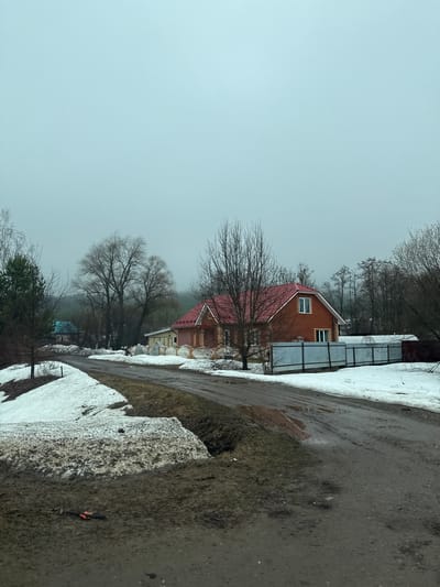 The image depicts a scene in Гремяча, Russia on an overcast day with visible snow accumulation. A brick house with a red roof stands prominently, partially obscured by bare trees. Adjacent to the house is a metal fence or structure. A road or path borders the scene, showing remnants of melted snow and muddy conditions. The sky is uniformly gray, suggesting a bleak or wintry atmosphere. In the foreground, piles of melting snow are visible along the roadside. The overall impression is one of a rural or semi-rural setting in late winter or early spring.