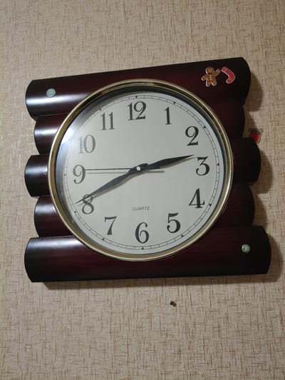 The image shows a dark brown, log-shaped wall clock mounted on a beige textured wallpaper. The clock face is white with black Arabic numerals and hands. The hour hand is pointing at approximately 8:40, and the minute hand is pointing at approximately 8:40. The clock has a gold-colored rim. There are small decorative Christmas-themed stickers on the clock's housing: a gingerbread man and a red and white stocking on the top right, and a red gift box on the right side. There are also two small green circular embellishments, one on the top left and one on the bottom right of the clock housing. A small screw or nail is visible in the wallpaper below the clock. The location is identified as the city of Chornobaivka, Ukraine.