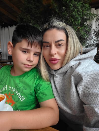 A close-up, indoor selfie shows a woman and a young boy posing for a photo. The woman, on the right, has blonde hair and is wearing a gray hooded sweatshirt. Her lips are painted a shade of pink. The boy, on the left, has dark hair and is wearing a bright green t-shirt with an orange graphic. He is leaning his head against the woman. In the background, a dark green coniferous tree is visible, with part of a wooden ceiling and a white curtain. A bowl of red soup is partially visible on the table in front of the boy. The scene appears to be set in a restaurant or cafe, possibly at the "Atoluka" restaurant in Atoluka, Bulgaria, given the presence of the distinctive tree and rustic ceiling.