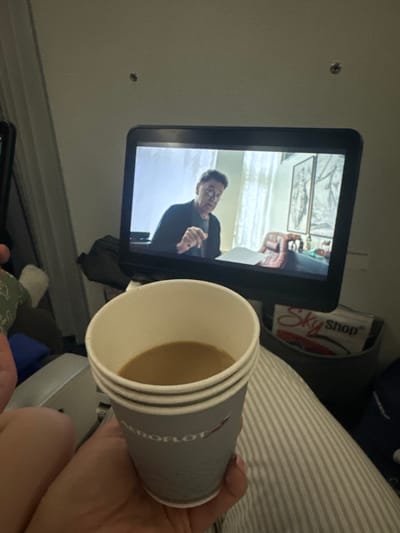 On an airplane, a passenger holds an Aeroflot paper cup filled with coffee in the foreground. In the background, an in-flight entertainment screen displays a man in a dark sweater, likely an actor in a movie. A Sky Shop advertisement is visible next to the screen. Striped fabric, possibly a blanket, is partially seen.