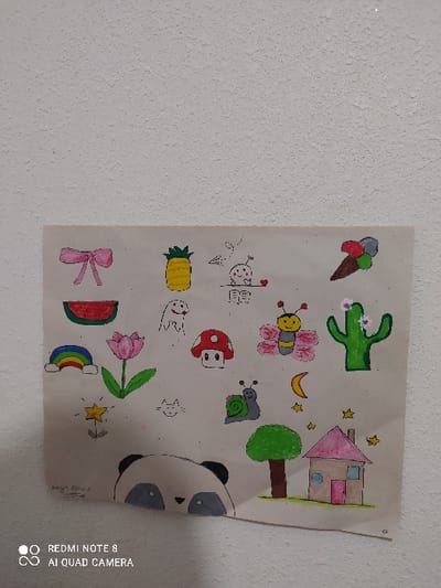 A drawing on beige paper depicts a variety of whimsical images. These include an ice cream cone, a cactus, a butterfly, a snail, a mushroom, a ghost, a flower, a watermelon, a rainbow, a star, a pineapple, a house, a moon, and a bow. Part of a panda's face is visible on the bottom right. The drawing appears to be affixed to a textured, light-colored wall.