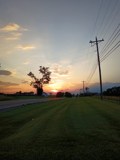 The image depicts a rural roadside scene at sunset in Lancaster, United States of America. The sky is a gradient of pale blue to orange, with scattered clouds tinged by the setting sun. A prominent power line pole stands to the right of the frame, with multiple wires extending across the sky. The foreground is dominated by a grassy embankment sloping down towards a paved road on the left. The road has a white dividing line. A silhouetted tree with a unique branch structure stands near the horizon, with a white house visible behind it. In the distance, a line of trees marks the edge of the landscape. The overall atmosphere is serene and tranquil, characteristic of a rural sunset.