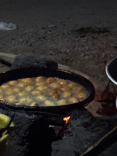 In the dim light of what appears to be nighttime, a large, black, cast-iron pot is positioned over a fire pit constructed with dark, rough stones. The pot is filled with what seems to be hot oil in which numerous round, golden-brown fritters are cooking. The fritters are submerged and bubbling, indicating they are actively being fried. Some of the fritters appear to have just been placed in the oil, while others are closer to being done. A large, dark, irregularly shaped object, possibly a cooking stone or a lid, is placed behind the pot. In the foreground to the left, partially visible green food items, possibly plantains, are present. The background is dark and out of focus, showing the rough texture of the ground, which is covered in small stones and dirt. A white piece of fabric or plastic is visible in the upper left corner of the image. The overall scene suggests an outdoor cooking setup in a rural or informal setting, potentially in the city of Angwan Sayawa, Nigeria, where traditional cooking methods over open fires are common.