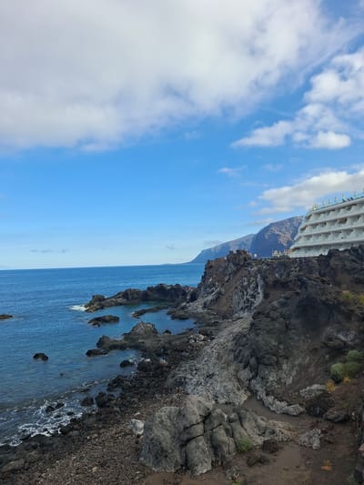 The image shows a coastal view of Puerto de Santiago, Spain. In the foreground, there is a rocky, dark-colored shoreline with the ocean meeting the land. The water is a deep blue, with some white foam where waves crash against the rocks. In the mid-ground, a modern, multi-story white hotel with balconies is visible, built on a cliff overlooking the sea. The hotel has rows of white lounge chairs on its upper levels. In the background, large, rugged mountains rise up from the coast, with a clear blue sky dotted with white clouds above. The overall scene depicts a resort area on a rocky coast.