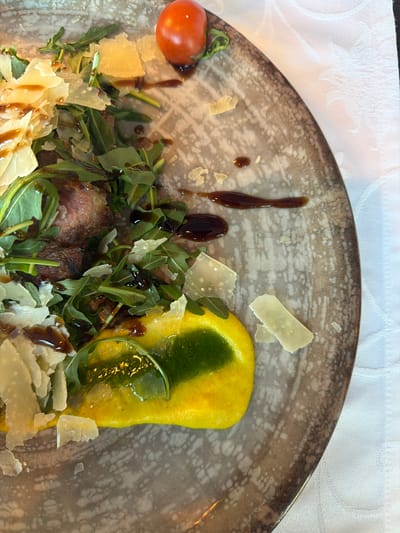 The image depicts a partially eaten plate of food, possibly a starter or a small main course. The plate is circular with a textured, mottled gray and brown pattern. The food elements consist of what appears to be grilled meat, possibly steak, mixed with arugula leaves and shavings of Parmesan cheese. A single cherry tomato is positioned on the left side of the plate. There is also a yellow, possibly polenta-based element topped with a green sauce. The dish is drizzled with a balsamic reduction. The plate sits on a white tablecloth with a subtle pattern. There is no visible indication in the image suggesting that this food is specifically from Grevenmacher, Luxembourg.