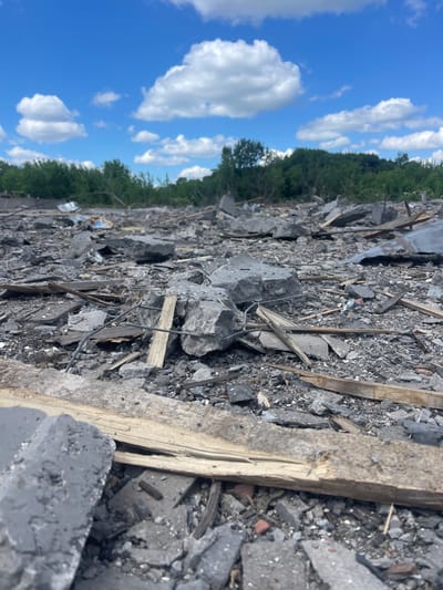 The image shows a field of rubble in Terny, Ukraine, at a location with coordinates of latitude 50.979 and longitude 33.946. The ground is covered in debris from a destroyed building, including broken concrete blocks, wood, and metal scraps. The sky above is a bright blue with white puffy clouds. In the background, there is a line of green trees. The scene suggests that this area has been subject to a bombing or other act of violence. The image appears to be taken from a low angle, looking across the rubble.
