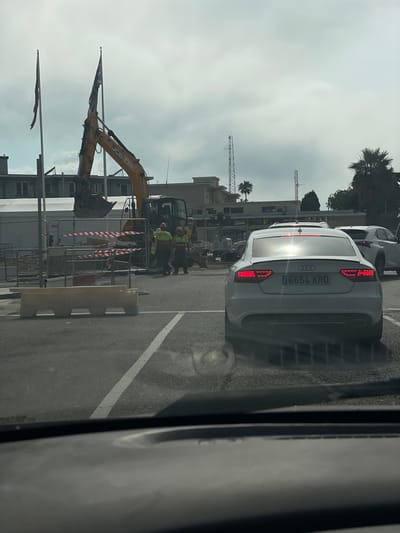 The image shows a street scene in La Línea de la Concepción, Spain. In the foreground, a white Audi A5 with license plate "6654 KRD" is parked. Behind it, construction is underway, with yellow excavators and several workers in high-visibility vests and hard hats. Barriers and caution tape are also visible. Two flags are mounted on poles, one with a red and white striped pattern and another with a darker color. In the background, buildings and palm trees suggest an urban or semi-urban environment. The sky is overcast, indicating a cloudy day. The lighting suggests it could be daytime. The overall scene depicts an active construction site next to a road where vehicles are parked.