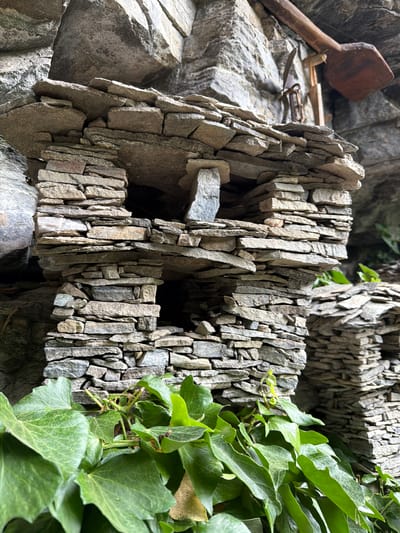 The image depicts a multi-tiered dry-stone structure, resembling miniature houses, constructed from numerous flat, irregularly shaped grey and brown stones stacked without mortar. The structure features at least two levels with open sections acting as windows or entrances, and is partially integrated into a larger, textured rock face in the background. Prominent green ivy with large, lobed leaves grows in the foreground and around the base of the stone construction, with additional green foliage visible further back. Above and to the right of the stone structure, a rustic, dark wooden object resembling a large paddle or scoop is affixed to the rock face, alongside a dark, aged metal object. The scene is set in a rocky, natural environment, specifically located in Golyamo Belovo, Bulgaria. No people are visible.