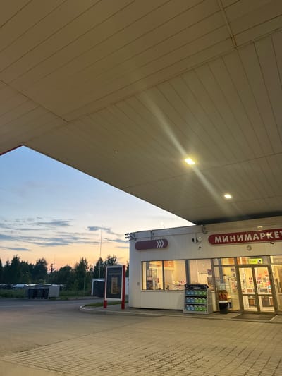 The image depicts the exterior of a minimarket located at a gas station, with a portion of the canopy and the sky visible. The minimarket has large windows and glass doors, with signs displaying "МИНИМАРКЕТ" in red lettering. Inside, shelves stocked with products are visible, and a display of cleaning supplies is positioned outside the entrance. In the background, trees and a cloudy sky at dusk or dawn are present. The ground is paved with interlocking bricks. The presence of trash bins, a portable toilet, and a sign with Cyrillic text suggest a location in Russia, likely in Krasnokamsk given the prompt. No people are clearly identifiable in the scene, though some indistinct figures may be present behind the glass windows. The scene is static and appears to be captured during twilight hours.