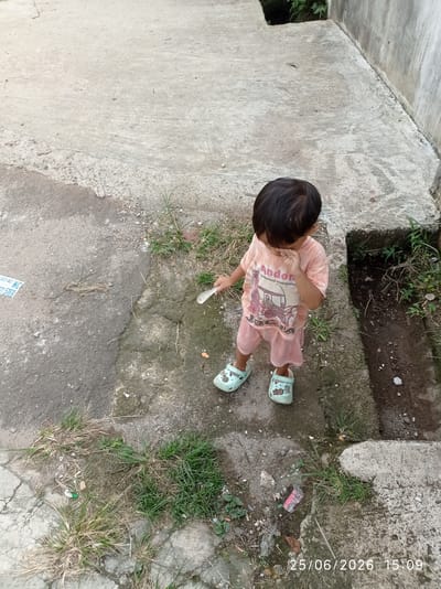A young child, likely a toddler, stands outdoors on a paved surface with some patches of grass. The child is wearing a pale pink t-shirt with a graphic print and matching pink shorts, along with light blue clogs. The child's hand is raised to their mouth. In their other hand, the child holds a white object that appears to be a spoon. The child is looking downwards and to their side.

The setting appears to be an outdoor area with concrete ground that is cracked and weathered in places. There are also clumps of dry and green grass growing through the cracks and along the edges of the pavement. To the right of the child, there is a darker, somewhat recessed area with soil and vegetation, possibly a drainage ditch or a garden bed. A concrete wall runs along the far right of the frame. The lighting suggests it is daytime, possibly late afternoon given the timestamp.

Visible text in the image includes a timestamp in the bottom right corner: "25/06/2026 15:09". There is also text and a graphic on the child's t-shirt, and a partially visible blue and white sticker or label on the ground to the left of the child. The text on the t-shirt appears to include "Andon" and "Traditional Transporta".
