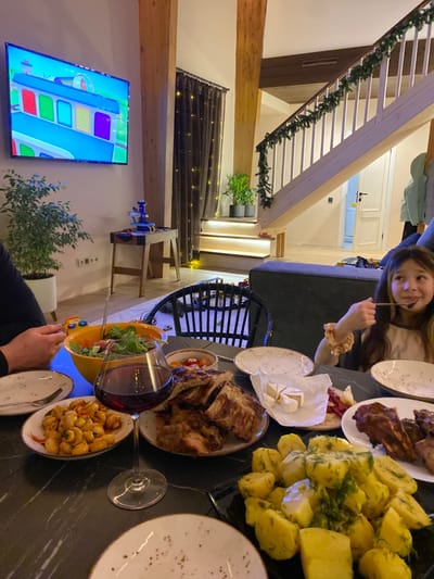 The image depicts an indoor dining scene occurring at a modern residential property in Burdakovka, Russia. The setting features light-toned wooden structural beams and a light-colored wooden staircase adorned with a green garland, indicating a possible holiday period. A large flat-screen television mounted on the left wall displays a colorful animated scene featuring a blue vehicle. Beneath the television, a small wooden console table holds a blue and white toy structure. Dark curtains with string lights are visible behind the console table, and potted plants are situated nearby and on a lighted step.

In the foreground, a dark-colored dining table is set with multiple dishes. Observable food items include a large plate of grilled ribs or meat, a plate of boiled potatoes with dill, a plate of white beans in a sauce, and a plate containing sliced cheese (likely Brie) and cured meat. A prominent orange bowl contains a green salad, and a wine glass filled with red wine stands on the table. Several empty or partially used white plates are also present.

Two individuals are partially visible. On the left, a person's arm and hand are near the salad bowl. On the right, a young girl with long, light brown hair, wearing a light-colored shirt, is shown from the chest up, holding a spoon to her mouth and appearing to be eating. She looks slightly off-camera with a subtle smile. A dark sofa is partially visible in the mid-ground on the right. The room is illuminated with warm artificial light.