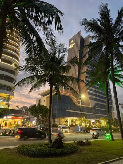 This image captures an urban street scene at dusk in An Hai Bac Ward, Son Tra District, Da Nang, Vietnam. The foreground features a landscaped median strip with green shrubs and a large dark rock. A multi-lane road is immediately beyond, with several vehicles in motion, including a dark SUV, a white van, and multiple motorcycles, some exhibiting motion blur. To the left, a rounded, multi-story building with illuminated horizontal bands stands beside a 7-ELEVEN convenience store. Several motorcycles are parked near the 7-ELEVEN, and indistinct figures are visible near its entrance. Dominating the right side of the frame is the Hotrans Hotel & Apartment, a tall, modern, angular high-rise building with glass and light-toned vertical paneling. Its exterior is brightly illuminated with decorative and functional lights, including a green vertical light strip on the far right and an illuminated "HOTRANS HOTEL & APARTMENT" sign near its peak. Numerous tall palm trees are prominent throughout the scene, some lit with green upward-facing lights, framing the buildings and partially obscuring the twilight sky, which displays hues of orange, pink, and grey. Streetlights are active, casting warm illumination on the road and surrounding structures.