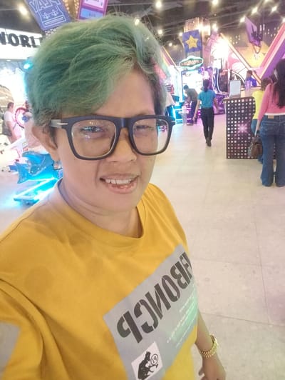 A medium shot shows a person with teal-colored hair wearing glasses and a yellow t-shirt in what appears to be an indoor arcade or amusement center. The person is looking at the camera and smiling. The t-shirt has a gray rectangular design with black text that reads "BONG" in large letters and some smaller text underneath. A gold-colored bracelet is visible on the person's left wrist. In the background, there are several arcade machines with colorful lights and various individuals walking around. One person in the background wears a pink top and blue jeans, another wears a yellow top and blue jeans, and a person in a blue shirt and black pants is walking away from the camera. The floor is tiled, and the overall lighting suggests a brightly lit, commercial indoor space. The location is identified as an arcade in Gunung Putri, Indonesia.