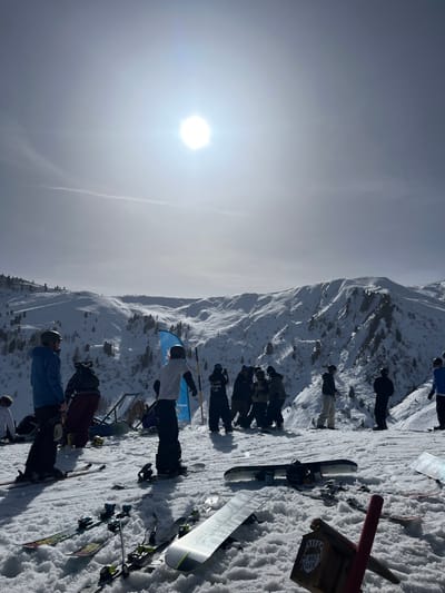 The image depicts a bright, sunlit winter mountain scene on a snow-covered slope in Schwendau, Austria, specifically within what is indicated by a partial sign as the "PENKEN PARK" area. The foreground consists of a groomed snow surface where multiple pairs of skis and several snowboards are laid out.

Approximately 15-20 individuals are visible across the scene, all dressed in winter sports attire, including jackets, pants, helmets, and goggles. On the left, a person wearing a blue jacket and helmet stands on skis, facing towards the right. Slightly to the right and foreground, another individual in a white t-shirt and dark pants, also wearing a helmet, stands on a snowboard. In the mid-ground, a group of approximately six people are clustered together, some appearing to be interacting, with one person holding a mobile phone. Behind this group, a blue and yellow banner or flag with partially visible white text (possibly "DECATHLON") stands vertically. Other objects include various portable loungers or chairs and some metal structures, consistent with a snow park, visible further up the slope. A wooden sign bearing the text "PENKEN PARK" and a bear paw logo is partially visible in the bottom right corner, alongside a red pole.

The background features expansive, snow-covered mountain ranges, with scattered coniferous trees visible on the slopes. The sky is clear and bright, dominated by the brilliant, high-contrast sun positioned in the upper center, which casts a strong halo effect and shows a faint contrail. No dynamic actions like skiing or snowboarding are observed; individuals appear to be resting or socializing.
