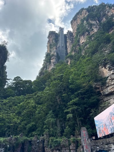 The image depicts a breathtaking natural landscape with imposing, lushly vegetated rock formations. Dominating the scene is a towering, glass-enclosed outdoor elevator affixed to the sheer face of a colossal cliff. The elevator, made of metal and glass, rises vertically, a stark contrast to the natural rock and dense foliage. The cliffs themselves are characterized by layered rock strata and are heavily overgrown with a variety of trees and vegetation, creating a vibrant green canopy that partially obscures the rock faces.

In the foreground, at the base of the cliffs, is a rocky outcrop with some man-made elements. There are tiered rock structures that appear to support small waterfalls, though they are partially obscured. The lower section of the image also shows a large, brightly lit LED screen displaying a scenic mountain landscape with vibrant colors. Adjacent to the screen, a tall, dark grey rock stands prominently, bearing large red Chinese characters. The sky above is partly cloudy, with a mix of grey and white clouds interspersed with patches of blue sky, suggesting an overcast but possibly brightening day. The overall impression is one of awe-inspiring natural beauty augmented by human engineering.