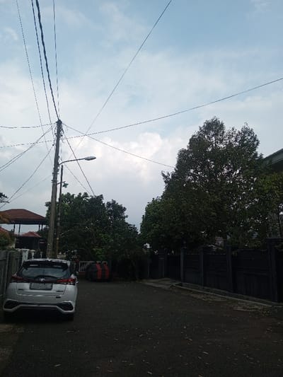 A street view showcases a white car parked on the left. Overhead, power lines crisscross against a cloudy sky. To the right, a dark fence lines the road, bordering a large tree. On the left, part of a gazebo roof is visible above the car. A street light is attached to a utility pole, adding to the urban ambiance. The road surface appears to be asphalt, showing signs of wear.