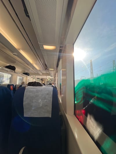This image captures the interior of a train carriage, purportedly in the city of Tortosa, Spain. The perspective is from a passenger seat, looking forward down the aisle.
**Environment:**
The train carriage features dark blue fabric seats, some equipped with white disposable headrest covers. The seat directly in front of the camera shows the back of a person's head with dark, braided hair. Further down the aisle, additional passengers are visible, mostly indistinct. The ceiling consists of white panels, overhead luggage racks holding several dark bags, and perforated ventilation grilles. Recessed lighting fixtures, both linear and rectangular, are illuminated, casting a warm glow.
**Exterior (through windows):**
On the right side of the carriage, large windows display a clear blue sky. A bright sun is prominently visible, creating significant glare and lens flares on the window surface. Railway infrastructure, including overhead catenary lines and support structures, can be seen beyond the windows. A distinct, vibrant green, wavy marking or reflection, resembling graffiti, covers a substantial portion of the lower right window, obscuring the external view. A red light reflection is also visible beneath this green marking.
**People, Subjects, Objects, Actions, Interactions:**
The primary subjects are passengers seated within the train carriage. No specific actions or interactions beyond being seated are discernible. Objects include the train's internal fixtures, passenger luggage on overhead racks, and the exterior railway infrastructure. The strong sun glare and the green marking on the window are significant visual elements.