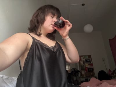The image depicts an indoor scene located in Cortona, Italy. A woman with short, dark brown hair is shown drinking from a clear, stemless glass filled with a dark red liquid, her eyes partially closed. She wears a black satin camisole with lace trim and a silver chain bracelet on her right wrist. The room is dimly lit, with a white spherical pendant light fixture visible overhead. In the background, a wall-mounted cork board with various items pinned to it is seen, along with a black standing fan and a red tapestry. Pink bedding occupies the lower right foreground.