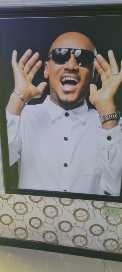 A framed picture of a bald man wearing sunglasses and a white shirt hangs on a wall with a circular pattern. The picture is rotated slightly clockwise. The man is smiling and holding his hands up. A plug socket is visible above the picture frame. The location is Alasia, Nigeria.