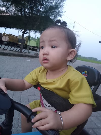 A young child is sitting on a tricycle, with their hands on the handlebars. They are wearing a yellow shirt and a silver bracelet. The child has dark hair tied up in a small ponytail. They are looking off to the side with a slightly pensive expression. The tricycle has a black seat and a blue frame. The background shows a paved area and an open space with a grassy field. There are some trucks parked under a covered structure. The photo was taken in Bungur, Indonesia.