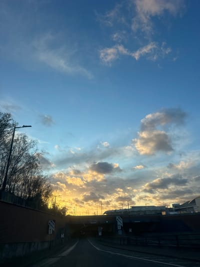 The image depicts a road approaching an underpass in Chatham, United Kingdom, specifically appearing to be an approach to the A2 (New Road) underpass near the Pentagon Shopping Centre. The scene is illuminated by either sunrise or sunset, evidenced by the low-angle golden light on the clouds and horizon.
The upper portion of the frame features a clear blue sky interspersed with scattered cumulus clouds, which are tinged golden and orange along their undersides towards the lower horizon.
In the foreground, an asphalt road with multiple lanes slopes gently downwards, appearing wet or reflective. White lane markings are visible. The road leads directly into a dark underpass or tunnel in the center of the lower frame.
On the left, a tall, dark brick retaining wall runs alongside the road, topped by a dense growth of bare deciduous trees silhouetted against the bright sky. A modern street light with a flat, rectangular head extends from the left margin above the trees. A white traffic sign with diagonal black stripes and an upward arrow is mounted on a pole near the wall, indicating lane direction, with a smaller blue 'P' parking sign beneath it.
On the right, above the underpass entrance, a dark, elevated structure spans the road. Above this, a large building complex with distinctive curved or wave-like rooflines is visible, appearing illuminated. Numerous street lights with similar flat, rectangular heads are positioned along this elevated structure and around the building. A green traffic light is visible above the underpass entrance on the right. Another white traffic sign, mirroring the one on the left, is present on the right, also with a blue 'P' parking sign. Faint text on an overhead sign above the underpass includes "CHATHAM."
The underpass itself is dark, with internal lighting or distant vehicle lights faintly discernible within. No individuals or specific vehicles are clearly visible in the immediate foreground.