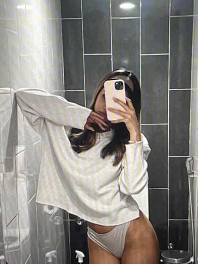 A young woman is taking a mirror selfie in a bathroom. She is wearing a white long-sleeved shirt and white underwear. Her left arm is bent at the elbow and resting on her head, while her right hand is holding a light pink smartphone up to take the picture. The bathroom features dark grey rectangular tiles on the back wall and lighter grey rectangular tiles on the side wall. A shower curtain is visible on the left side of the image. The image does not provide any information about the city of Pruszcz Gdański, Poland.
