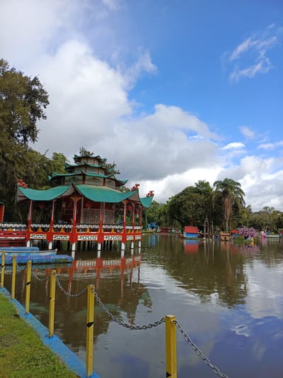 The image depicts a Chinese-style pavilion situated on a lake in the city of El Cisne, Ecuador. The pavilion has a green multi-tiered roof and red supports. It is surrounded by a body of water that reflects the structure and the sky above, which is partly cloudy with patches of blue. The foreground features a yellow and blue fence line. Beyond the pavilion, other structures and trees dot the landscape around the lake. The photo has good lighting and clarity.
