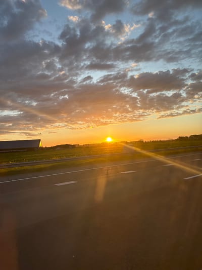 The image shows a landscape at sunset. The location is Beesd, Netherlands. To the left are dense, grey, and white clouds. The sun is setting on the horizon, creating a bright, golden light in the sky. To the right is a straight, empty road with white lines. There is a grassy area beside the road. The sun's reflection is visible on the road surface.