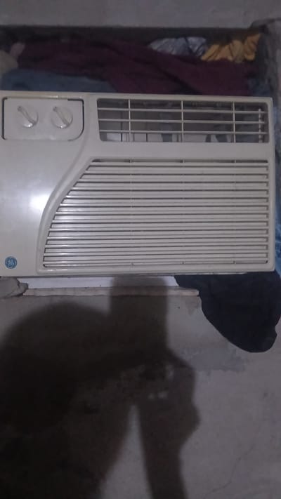 The image shows a GE window air conditioner resting on the floor against a wall. There is bedding and fabric on the left side of the unit, and a dark piece of cloth behind the air conditioner. There is a wooden shim next to the unit on the right. The image does not contain sufficient information to confirm its location within Putucual, Venezuela.