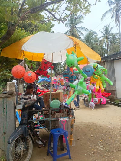 An outdoor scene in Meunasah Raya, Indonesia, features a young person sitting on a parked motorcycle, looking down at something in their hands. The motorcycle is laden with various items, including a wicker basket filled with goods and a blue plastic stool. Above, a yellow and white umbrella provides shade, from which several inflatable toys, including green shark-like creatures and a blue smiley face balloon, are suspended. In the background, palm trees and a building are visible, and other vendors and their stalls can be seen along a dirt road. Two orange basketballs are also hanging nearby, enclosed in mesh bags. The overall atmosphere suggests a bustling marketplace.