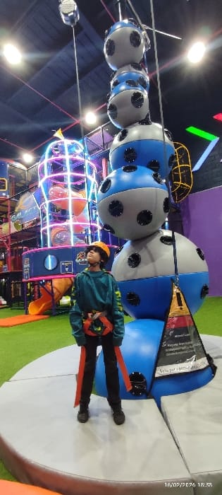 A child in a teal jacket and orange helmet, wearing a harness with orange straps, stands on a white and green cushioned surface. Overhead, a line of blue and gray spheres suspended from cables stretches across the scene. Below, a colorful play structure with tunnels and lights is visible. The date "16/02/2026" is stamped in the corner. The setting appears to be an indoor playground or amusement center.
