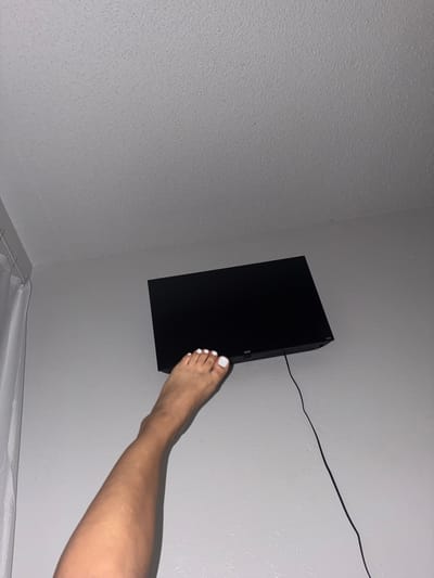 The image shows a close-up view of a person's leg and foot, with painted white toenails, extended towards a black flat-screen television mounted on a light gray, textured wall. A black power cord is visible trailing from the TV. The setting appears to be indoors, likely a bedroom or living area, given the presence of a shower curtain rod and curtain visible at the bottom of the frame. The lighting suggests it might be daytime, with artificial light reflecting off the wall and ceiling. The overall mood is casual and somewhat playful, with the person's leg positioned in a way that suggests interaction with the TV. The location context provided is Balch Springs, United States of America. No visible text is present on the TV or surrounding wall.