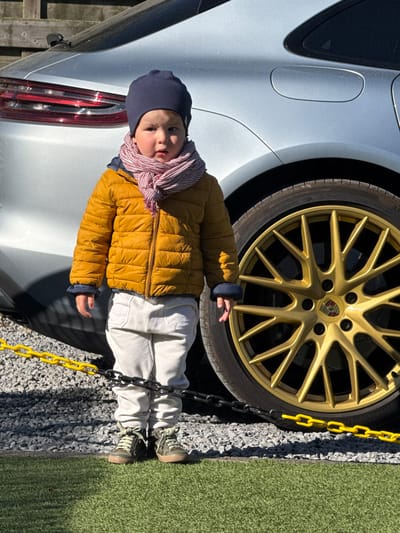 A young child stands outdoors in Bredene, Belgium, next to a silver car with distinctive golden wheels. The child, dressed in a dark blue beanie, a striped scarf, a mustard yellow puffer jacket, light-colored trousers, and dark green sneakers, looks towards the camera with a neutral expression.
The car, identifiable as a Porsche by the crest on its golden multi-spoke wheel, features a metallic silver finish. Text such as "OUTSIDE" is visible on the tire sidewall. A bright yellow plastic chain and a darker metal chain run along the ground, creating a low barrier between the child and a path of grey and black gravel. To the right of the gravel, a section of vibrant green artificial grass is visible. A wooden fence can be seen in the background behind the car's rear section. The scene is brightly illuminated by sunlight, indicating a clear, sunny daytime.