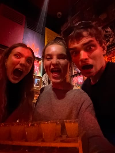 The media file is a low-light, vertically oriented selfie featuring three individuals with exaggerated expressions. Two women and one person with short, curly hair are positioned close together, their faces filling most of the frame. All three have their mouths open, as if shouting or reacting with surprise. The lighting is predominantly red, suggesting a dimly lit environment, possibly a bar or club. In the background, there are framed pictures on the wall, some with ornate gold frames, and what appears to be a ventilation grate. To the right, a wooden structure with multiple compartments, possibly a display case or shelving, is visible. The presence of what seems to be alcohol in some of the compartments is hinted at. The overall mood is energetic and spontaneous. Given the context of Jing'an District, China, this could be a snapshot of friends enjoying a night out. No visible text is present in the image.