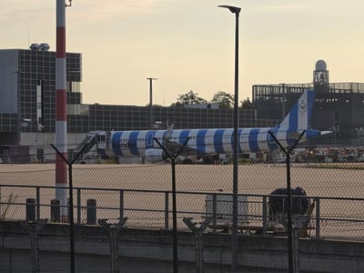 The image captures an airport scene at dusk, likely Frankfurt Airport, located in Kelsterbach, Germany. A Condor airline airplane, with a distinctive blue and white striped livery, is parked on the tarmac. The plane's tail features the Condor logo. In the background, airport buildings are visible, including a multi-story structure with a grid-like facade and a radar dome on top of another building. A red and white striped striped striped striped pole stands in the foreground, next to a lamppost. A chain-link fence with barbed wire runs across the bottom of the image, partially obscuring the view of the tarmac. The sky is a pale yellow, indicating the setting sun. Several airport service vehicles and equipment are scattered across the tarmac.