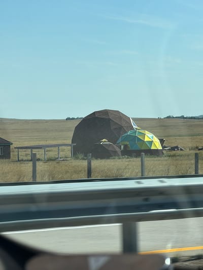 The image displays a landscape under a clear, pale blue sky. In the middle ground, there are several geodesic dome structures. One prominent dome is dark brown and appears to be covered in asphalt shingles. Adjacent to it is a smaller dome with a colorful, segmented facade, featuring panels in shades of yellow and teal. A smaller, dark brown dome structure is also visible to the left of the colorful dome. To the far left, a portion of a building with a dark roof and a white window is visible. Between the building and the domes is a covered, open-air structure with a multi-pitched roof supported by white posts. The ground is covered in dry, golden-brown grass. A metal guardrail runs across the foreground of the image, with a road visible behind it, blurred due to motion. The scene suggests a rural or semi-rural setting, possibly an isolated property. There are no people visible in the image. The location of Ereymentaw, Kazakhstan is not indicated by any visual cues within the provided image.
