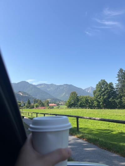 The image displays a sunny daytime scene in Fischbachau, Germany. In the immediate foreground, a human hand holds a white disposable coffee cup with a white lid, observed from within a vehicle; a dark vehicle frame is visible on the left. The midground features a paved path or road running parallel to a wooden fence, beyond which lies a large, vibrant green field. Scattered trees, including both deciduous and coniferous types, are visible behind the field. Several buildings with reddish-brown roofs are discernible in the distance, indicating a small settlement. A utility pole stands on the left side of the path. The background is dominated by a range of tree-covered mountains under a clear, bright blue sky with minimal, wispy white cloud formations. The scene is well-lit by natural sunlight, casting visible shadows.