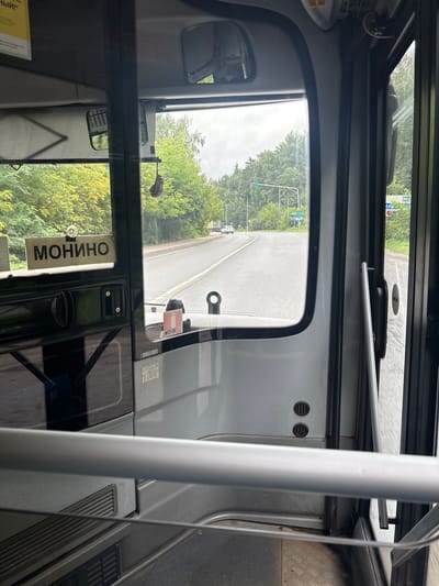 The image depicts the interior of a bus, looking forward through the windshield and side window. A sign with the Cyrillic word "МОНИНО" is visible on the dashboard. Outside, a road with trees lining both sides is seen. The road appears to be in a semi-rural or suburban area. There are traffic lights and other vehicles visible in the distance. Based on the sign "МОНИНО", this scene is likely located near Monino, which is a settlement in the Shchyolkovsky District of Moscow Oblast, Russia. The environment appears to be outdoors on a cloudy day, with lush green foliage indicating a temperate climate, likely summer or early autumn.